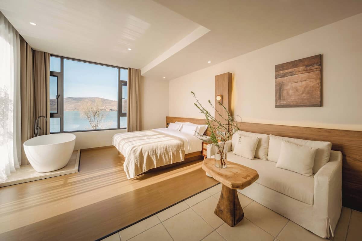 Une chambre d’hôtel moderne avec un grand lit, un canapé, une table basse en bois et une vue sur un lac.
