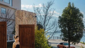 A woman in a black dress walking through a wooden door, with a stone wall, a tree, and a car parked on a street with a view of the sea. - Dali Antu Yuanchu seaview Homestay (Dali)
