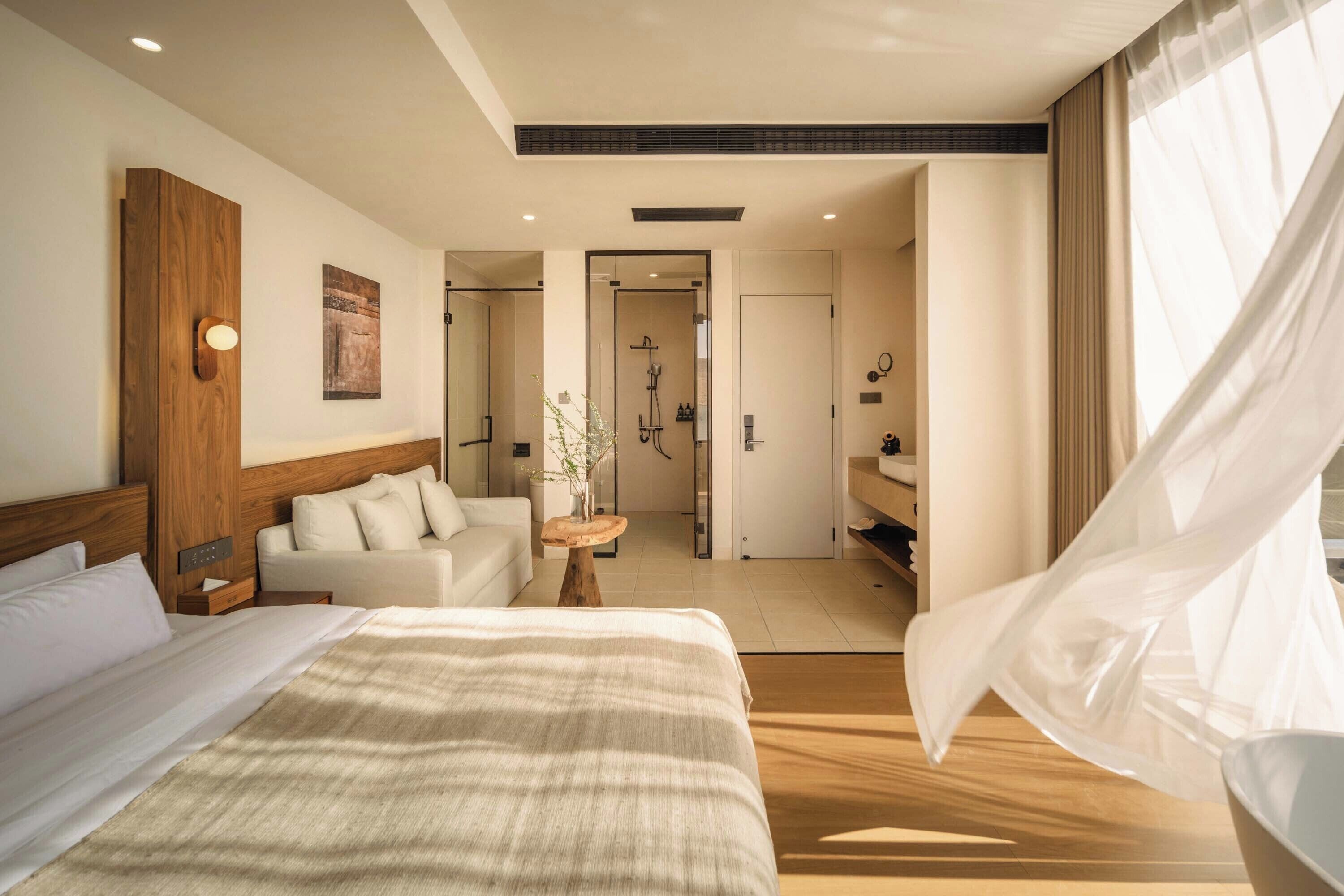 Habitación de hotel moderna con una cama grande, un área de descanso con sofá, una pequeña mesa con un florero y un baño visible a través de una puerta abierta.