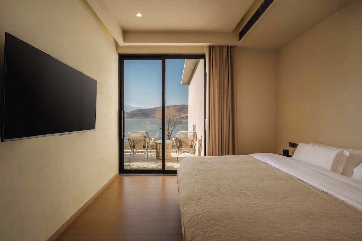 Une chambre d’hôtel dotée d’un grand lit, d’un téléviseur à écran plat fixé au mur et d’une porte-fenêtre coulissante donnant sur un espace extérieur aménagé avec des chaises.