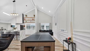 A spacious living room with a ping pong table, a black leather sofa, and a dining area with a kitchen in the background. - Upscale Retreat-Games, Comfort & Cozy Vibes Await! (Houston)