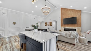 A modern kitchen with a marble island, a dining area with a sofa, and a living room with a TV and fireplace. - Upscale Retreat-Games, Comfort & Cozy Vibes Await! (Houston)