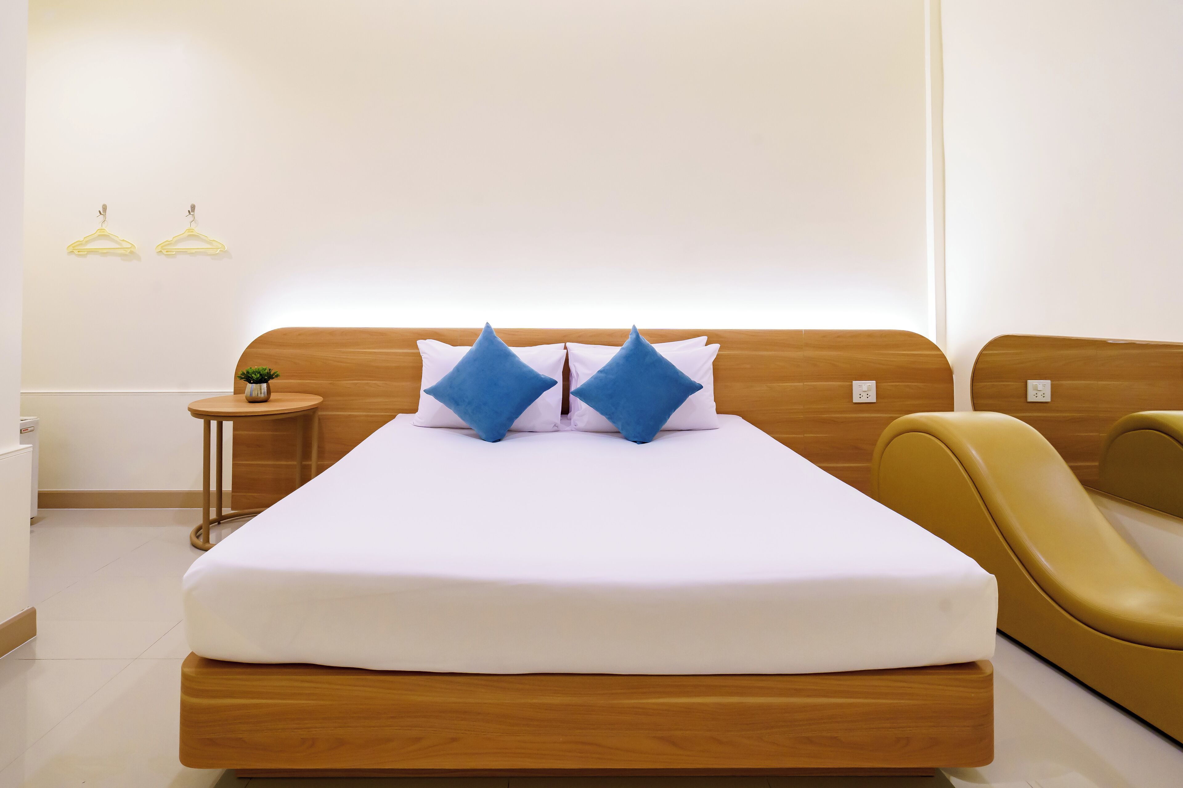 Habitación de hotel moderna con una cama de madera, dos almohadas azules, una mesita auxiliar con una planta y una silla amarilla.
