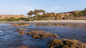 Una playa con formaciones rocosas, palmeras y un edificio al fondo.