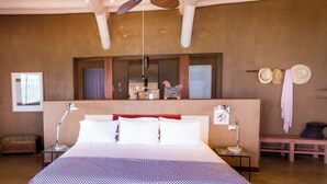 Un dormitorio amplio con una cama grande, un ventilador de techo, una mesita de noche con lámpara, un banco y un perchero en la pared.