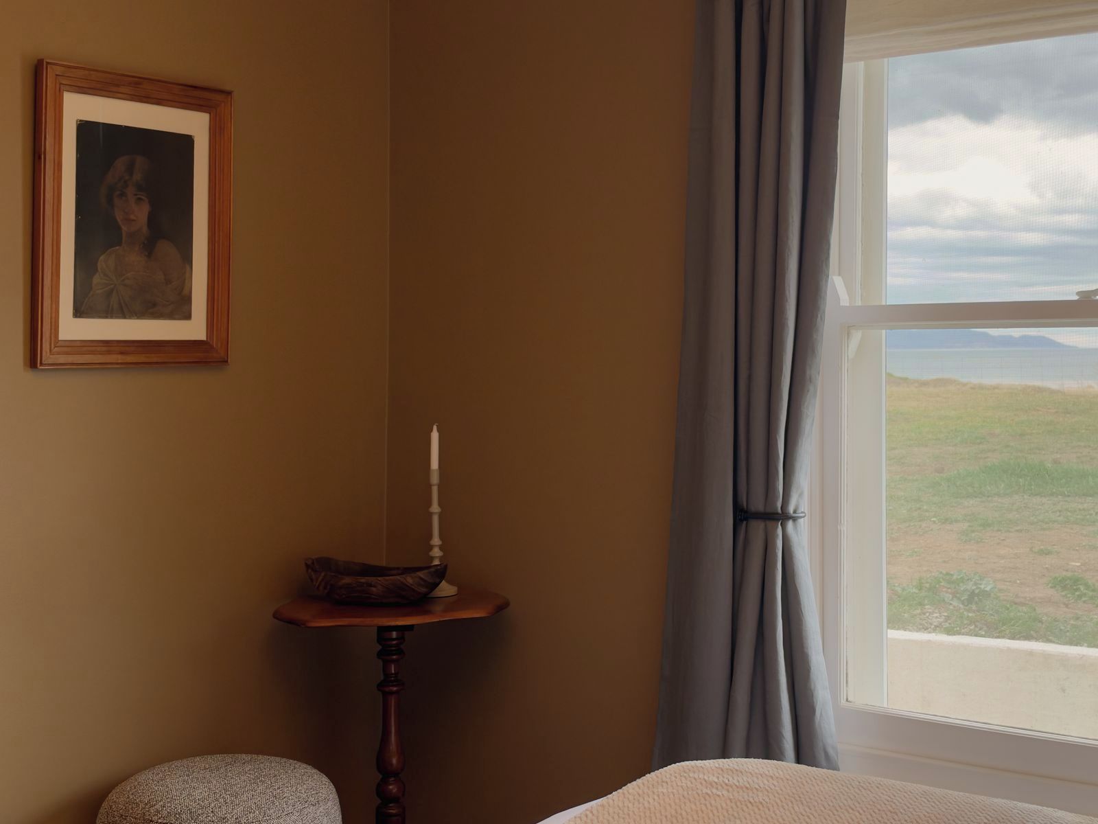 A room with a bed, a small table with a candle, a framed picture on the wall, and a window with a view of the landscape.