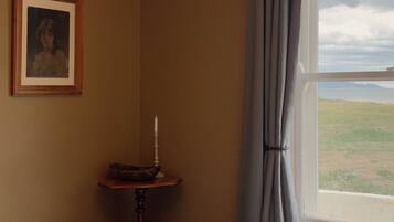 A room with a bed, a small table with a candle, a framed picture on the wall, and a window with a view of the landscape.