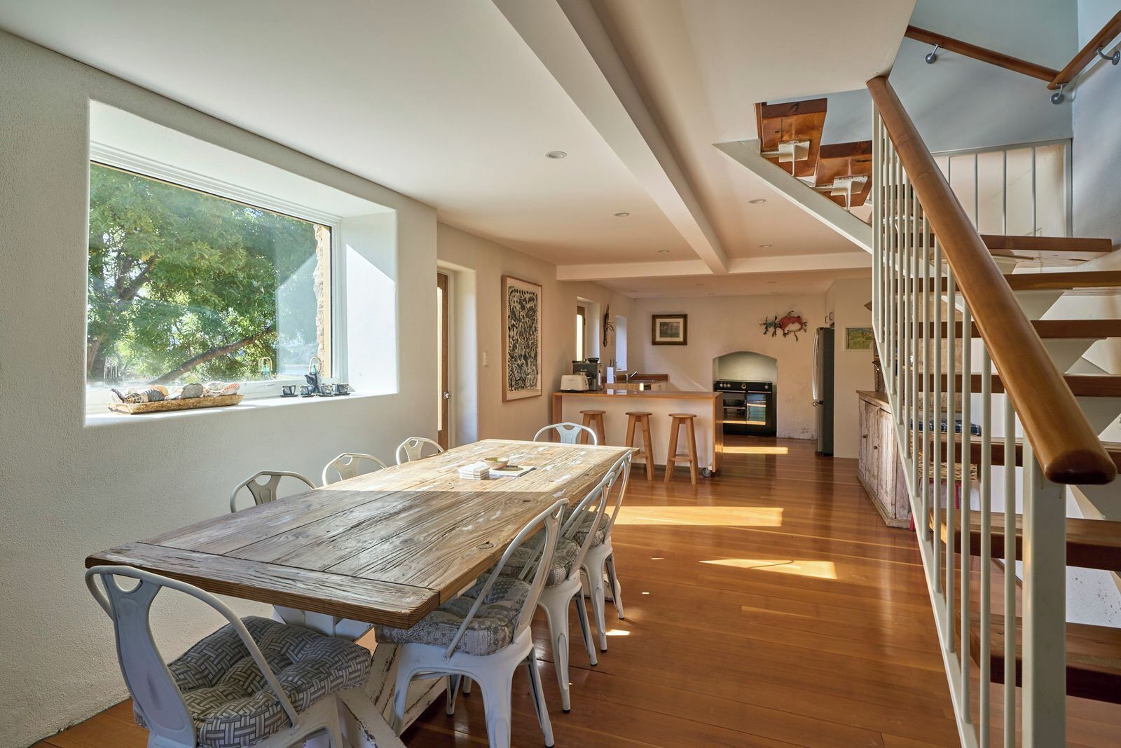 A spacious dining area with a long wooden table and white chairs, a kitchen in the background, and a wooden staircase.