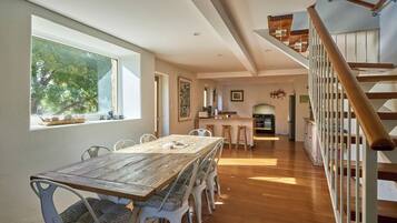 A spacious dining area with a long wooden table and white chairs, a kitchen in the background, and a wooden staircase.
