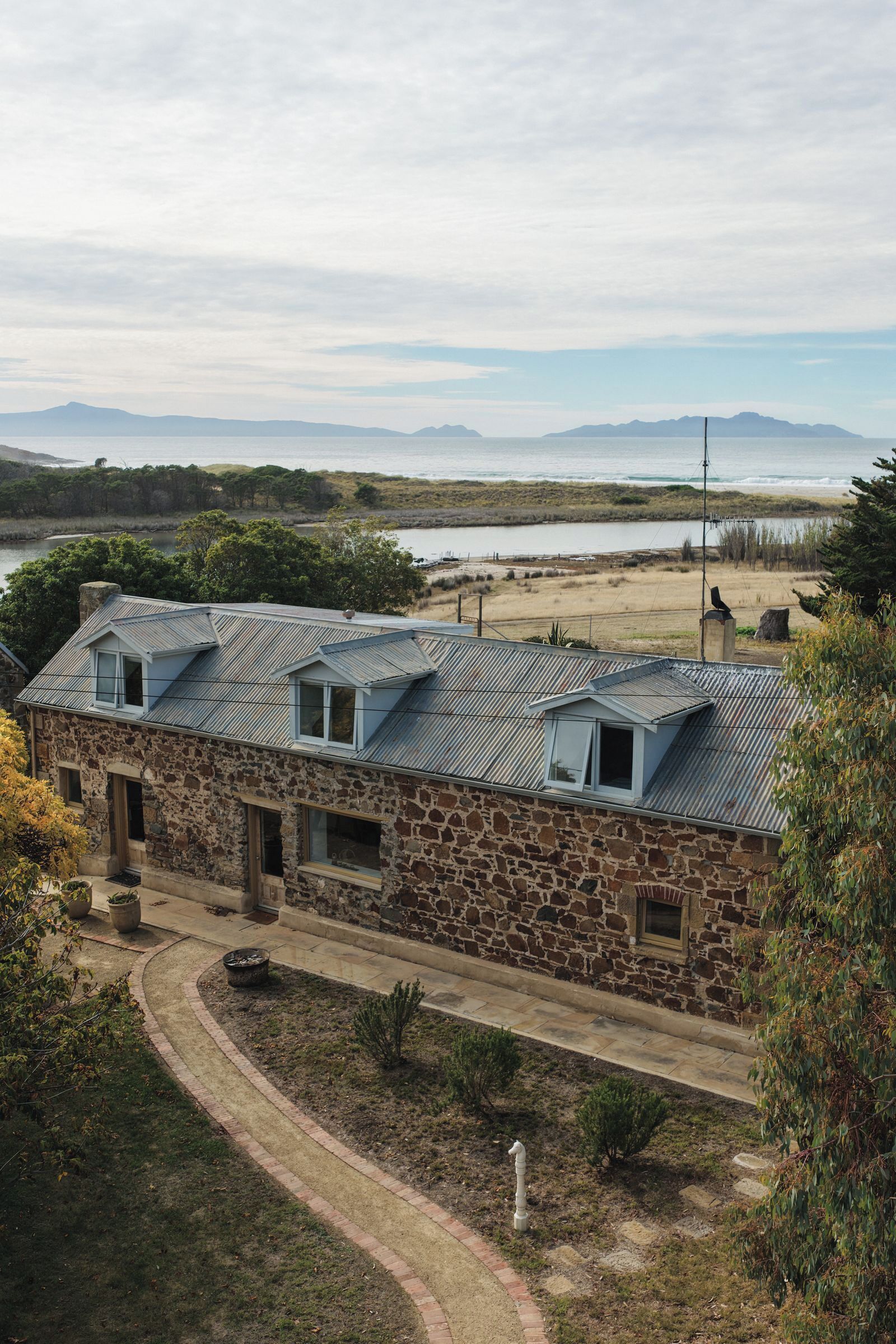 A stone building with a metal roof, a paved driveway, and a view of a body of water and mountains in the distance.