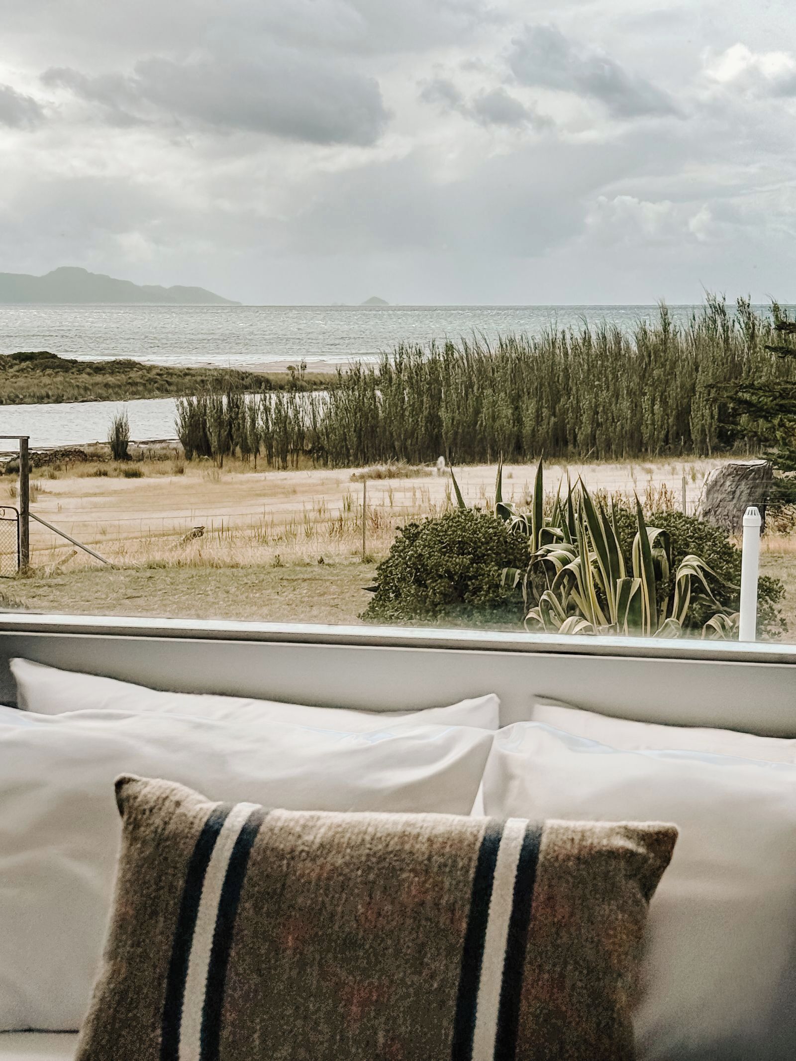 A view from a window with a sofa and a cushion, overlooking a beach with grass and a fence.