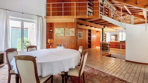 Un'ampia stanza con soffitto alto, travi in legno, una zona pranzo con tavolo e sedie rotonde, una scala e una biblioteca con scaffali.