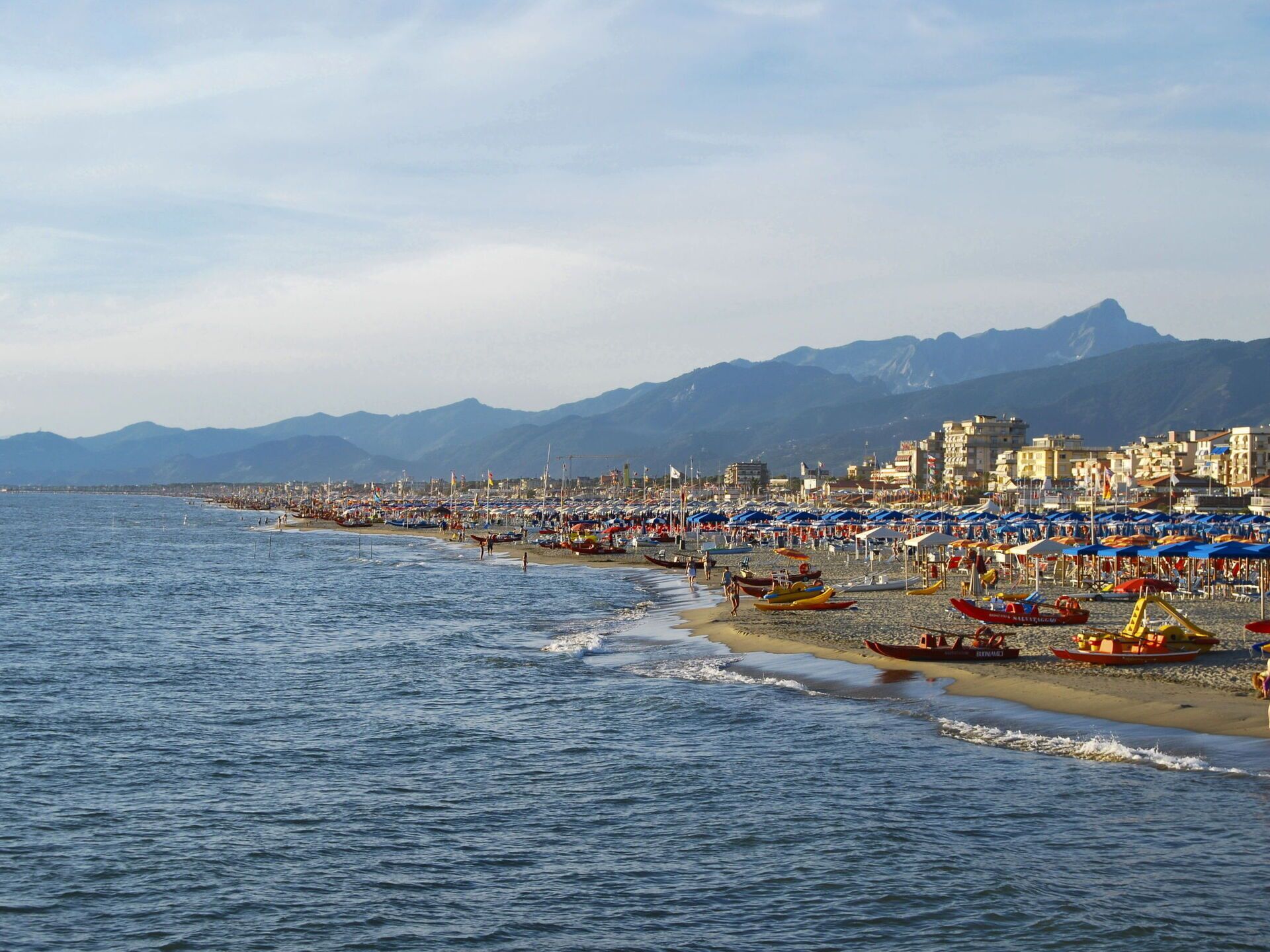 Una spiaggia costiera con numerosi ombrelloni e barche, cielo sereno e montagne sullo sfondo.
