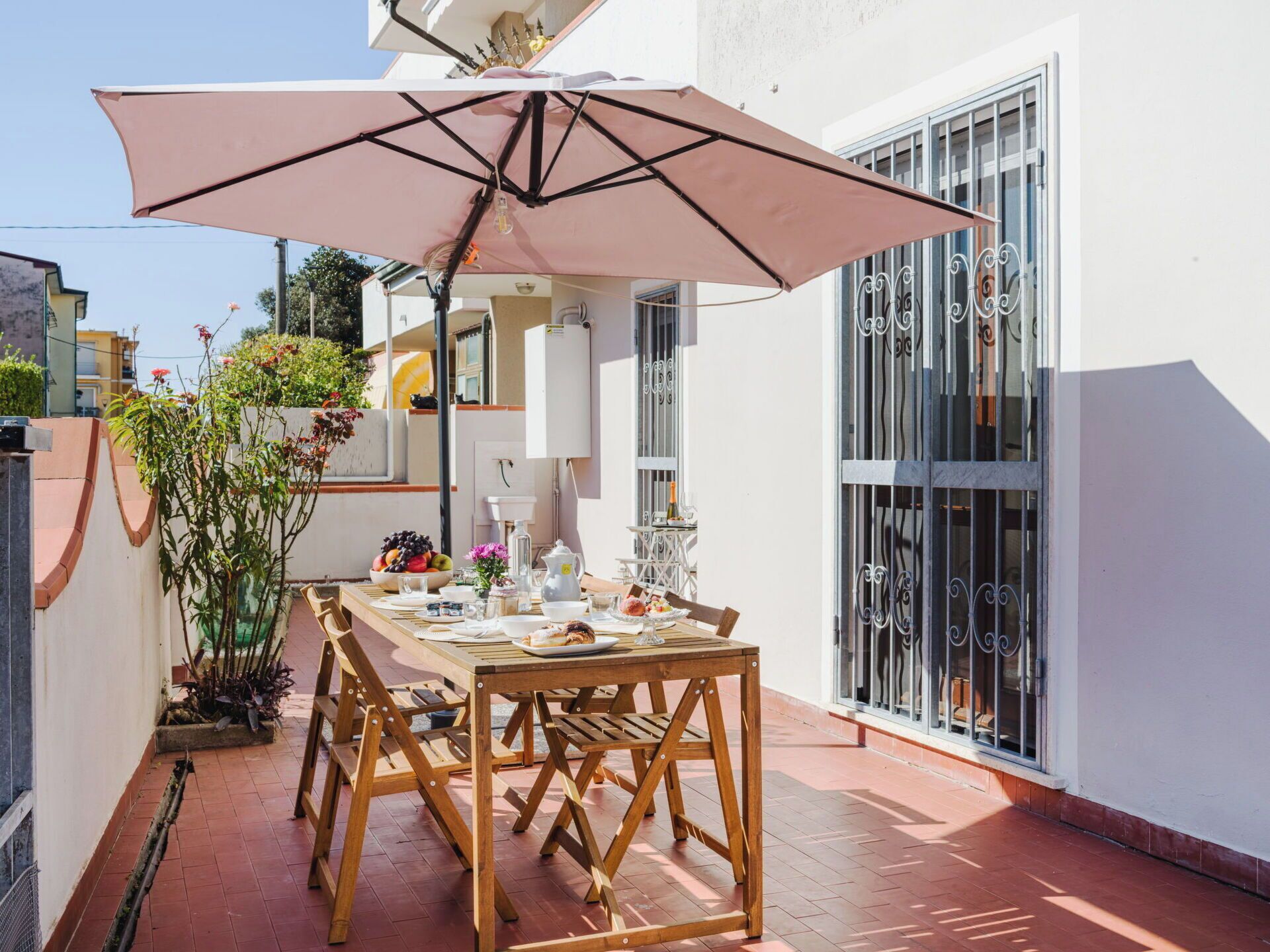 Una terrazza con tavolo apparecchiato per mangiare, circondata da piante in vaso e un ombrellone grande.
