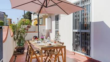 Una terrazza con tavolo apparecchiato per mangiare, circondata da piante in vaso e un ombrellone grande.