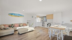 A modern living room with a sofa, a dining table with chairs, and a kitchen area with white cabinets and a window with blinds. - Seahaven Views in Ventnor (Ventnor)