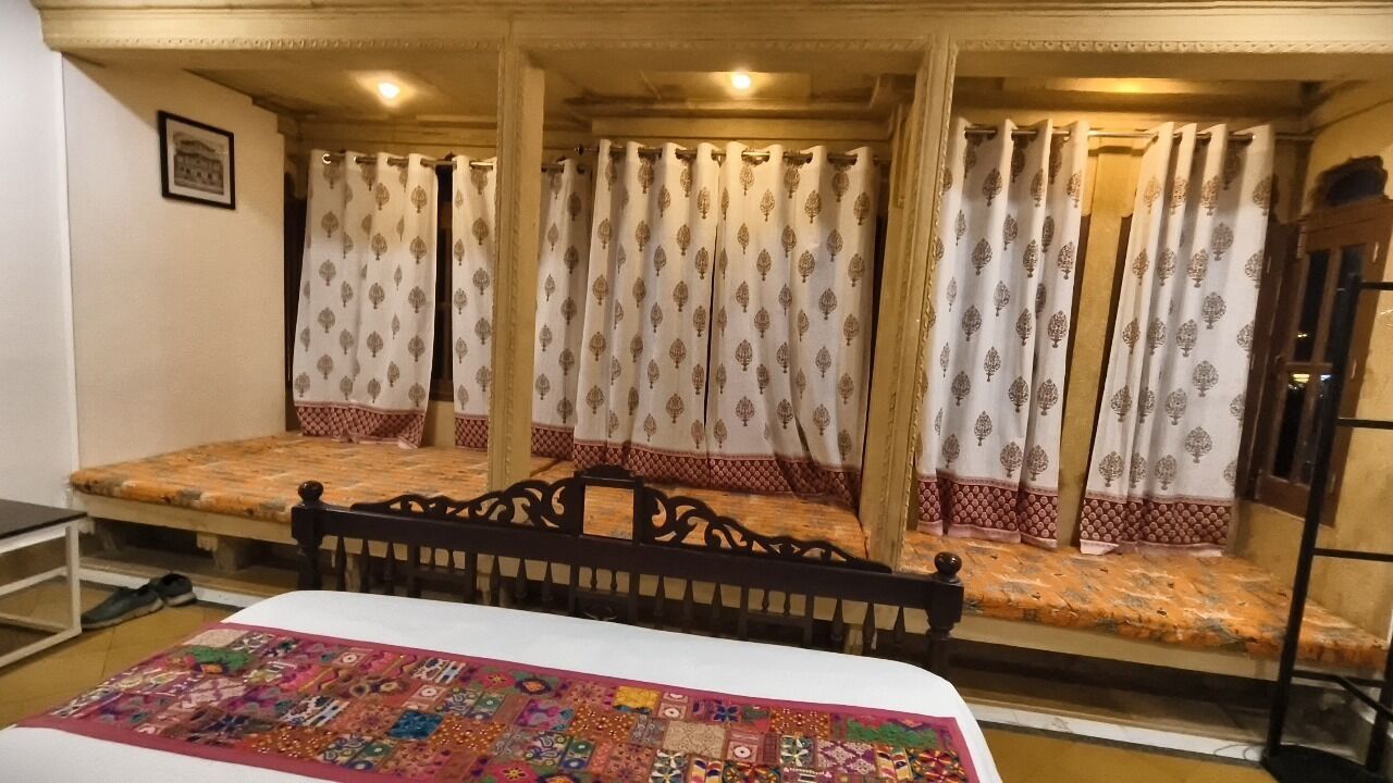 Ein Zimmer mit einem Bett, einer hölzernen Sitzbank und einem Fenster mit gemusterten Vorhängen.