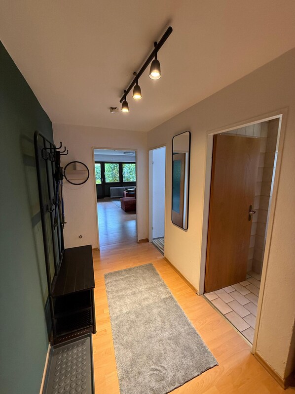 A hallway with a wooden floor, a grey rug, a black bench, a mirror, and a door leading to another room.