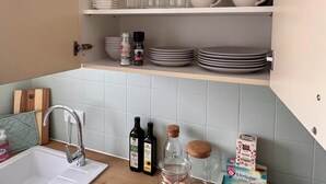 Una cocina con un lavamanos integrado, una encimera y repisas superiores que guardan platos, tazas y una caja con la etiqueta 'TEA'.