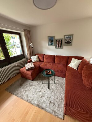 A modern living room with a sectional sofa, a small side table, and a radiator. There are two framed pictures on the wall and a window with curtains.