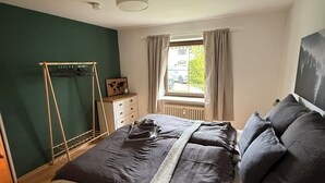 Un dormitorio con cama, cómoda de madera, perchero, ventana con cortinas y radiador.