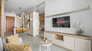 Un salon moderne avec un téléviseur fixé au mur, un canapé, une petite table avec une plante, et un coin repas à l’arrière-plan.