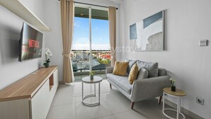 Un salon moderne avec un canapé gris, une console en bois, un téléviseur et un balcon agrémenté de rideaux.