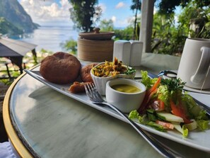 A table with a meal consisting of bread, a salad, soup, and a cup, set against a scenic view.