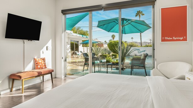 Une chambre d’hôtel avec un lit, un banc, une télévision et une vue sur un espace extérieur agrémenté de parasols et de palmiers.
