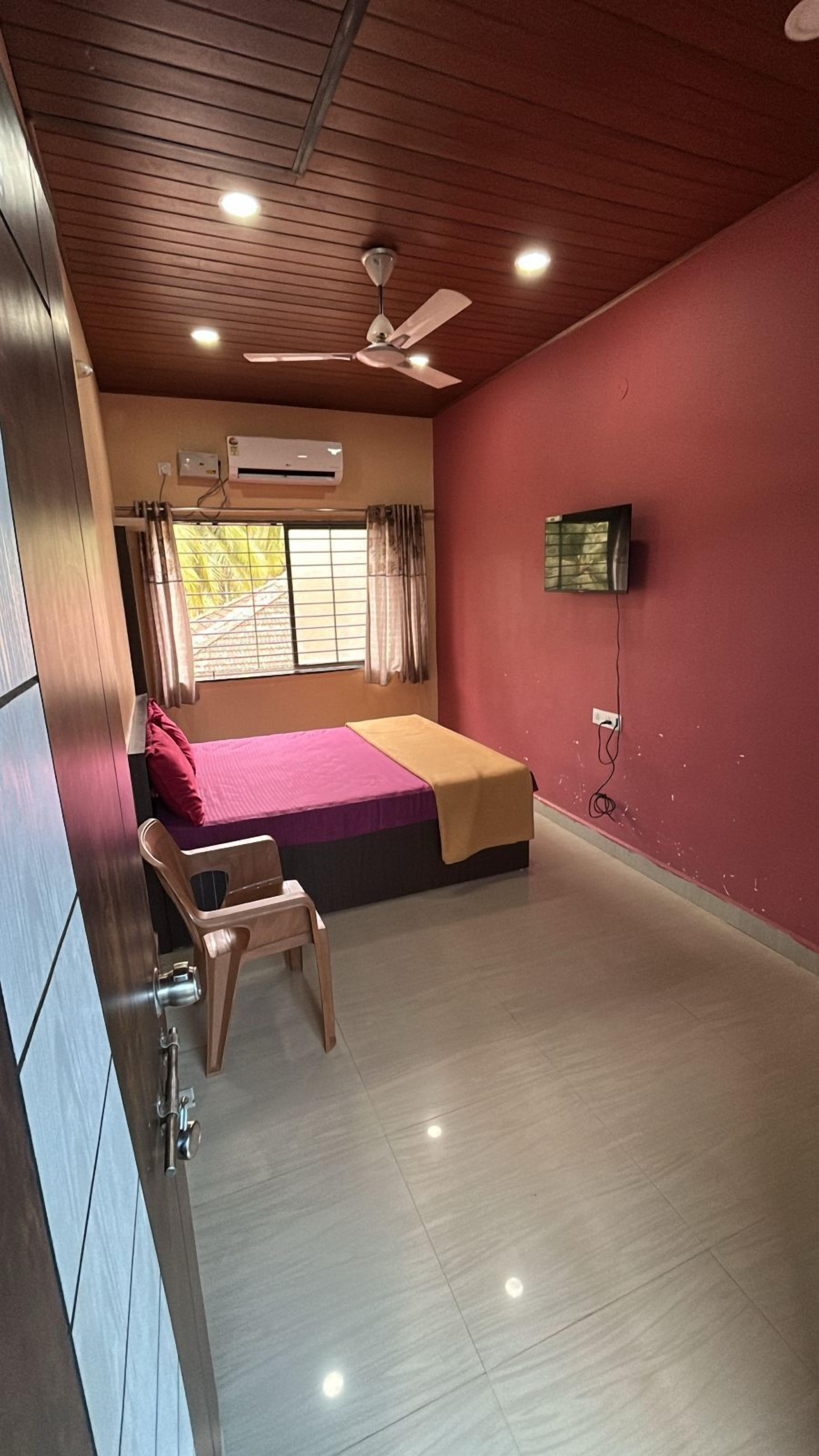 Quarto com cama, poltrona, ar-condicionado, ventilador de teto e televisão na parede.