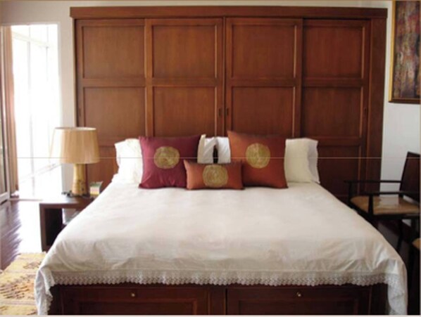 A large bed with a wooden headboard, a bedside table with a lamp, and a chair in a room with a wooden floor and a patterned rug.