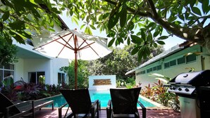 A deck with two chairs and a grill, overlooking a swimming pool and lush greenery. - Nakara Samui S4 Two Bedrooms Apt (Ko Samui District)