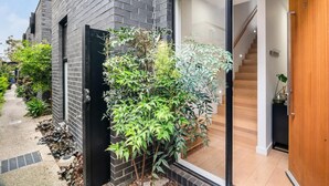 A modern house with a black brick exterior, a glass door entrance, a wooden staircase inside, and a small garden with various plants. - Chic Home with BBQ & Outdoor Dining (Carnegie)