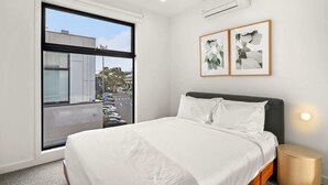 A hotel room with a large window, a bed, a bedside table, and two framed pictures on the wall. - Chic Home with BBQ & Outdoor Dining (Carnegie)