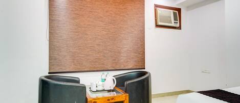 A room with a bed, a desk with a chair, a wall-mounted air conditioner, and a window with a brown blind.