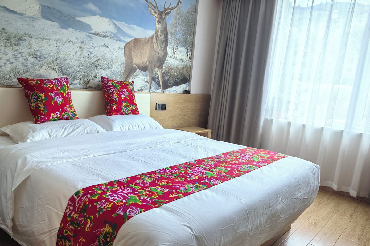 Habitación de hotel con una cama grande, ropa blanca, colcha de cama con motivos florales coloridos y un mural de un ciervo en la pared.