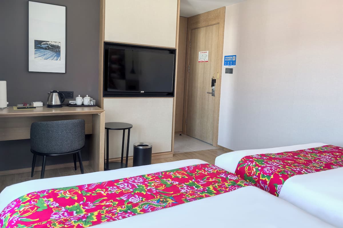 Habitación de hotel con escritorio, silla, dos camas con cubrecamas coloridos, televisor y una puerta con un letrero.