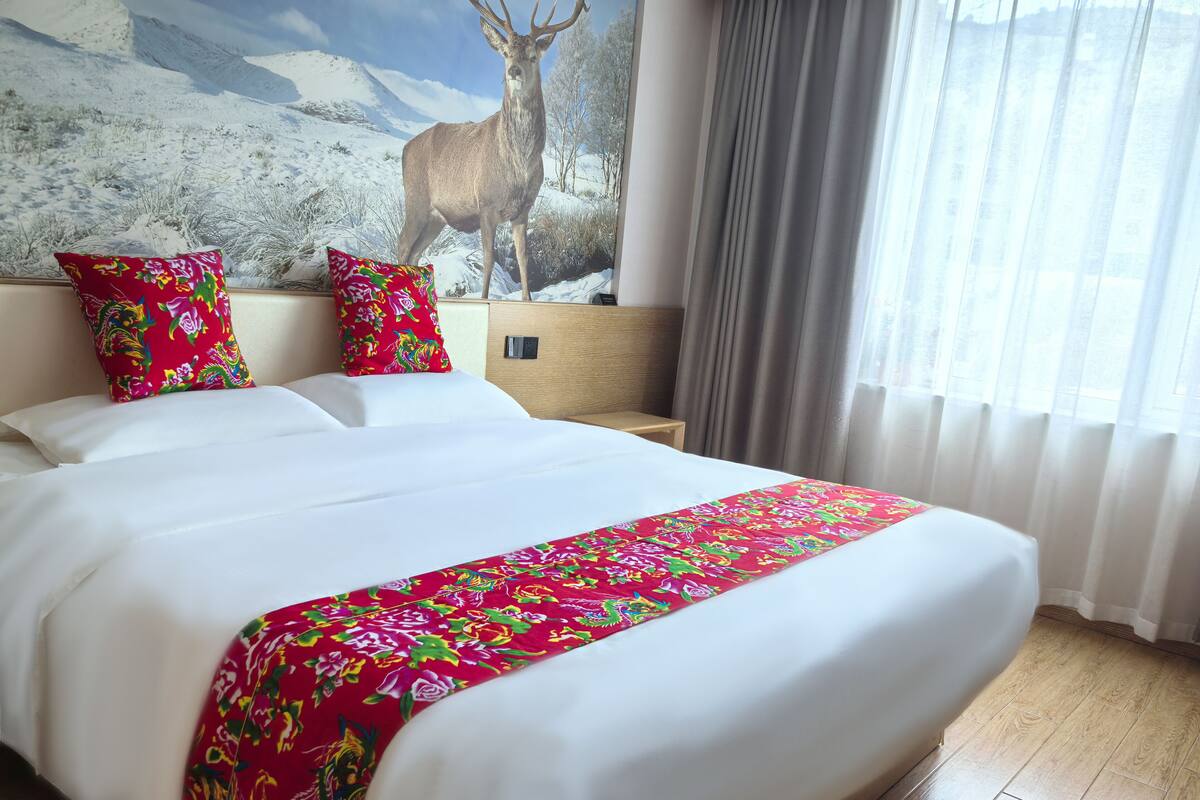 Habitación de hotel con una cama grande, cabecera de madera, colcha floral de colores y un mural de paisaje montañoso en la pared.