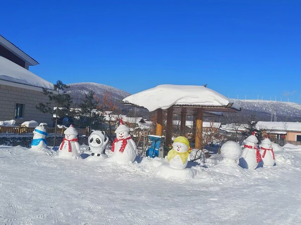 A snowman display in a snowy outdoor area with a clear blue sky and a wooden gazebo in the background.