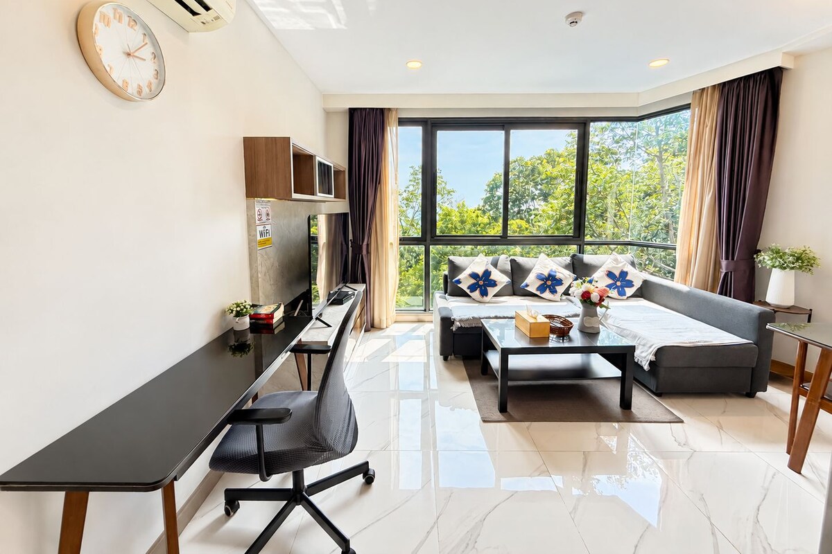 A modern living room with a sofa, coffee table, and a desk with a chair. There is a large window with curtains, a clock on the wall, and an air conditioning unit.