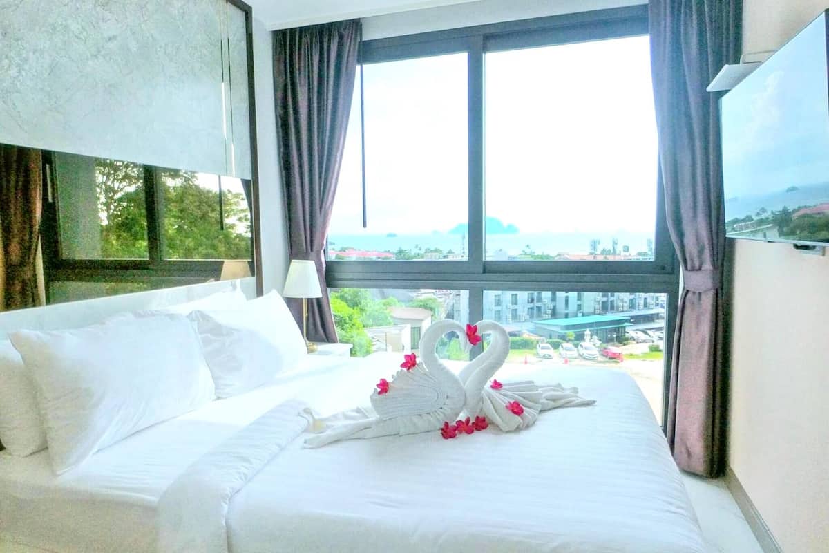 A hotel room with a bed, a window with curtains, and a view of buildings and greenery outside.