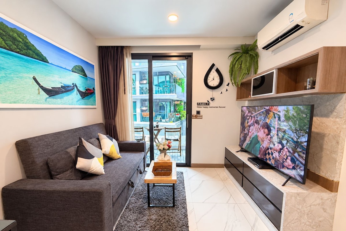 Una sala de estar moderna con un sofá gris, un televisor de pantalla plana y un ventanal con vistas a un edificio y zonas verdes.
