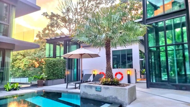 A modern outdoor pool area with a palm tree, lounge chairs, and a glass building in the background. - Silk Ao Nang Condominium (Krabi)