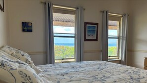 Un dormitorio con cama, dos ventanas con cortinas, un cuadro en la pared y vista al mar.