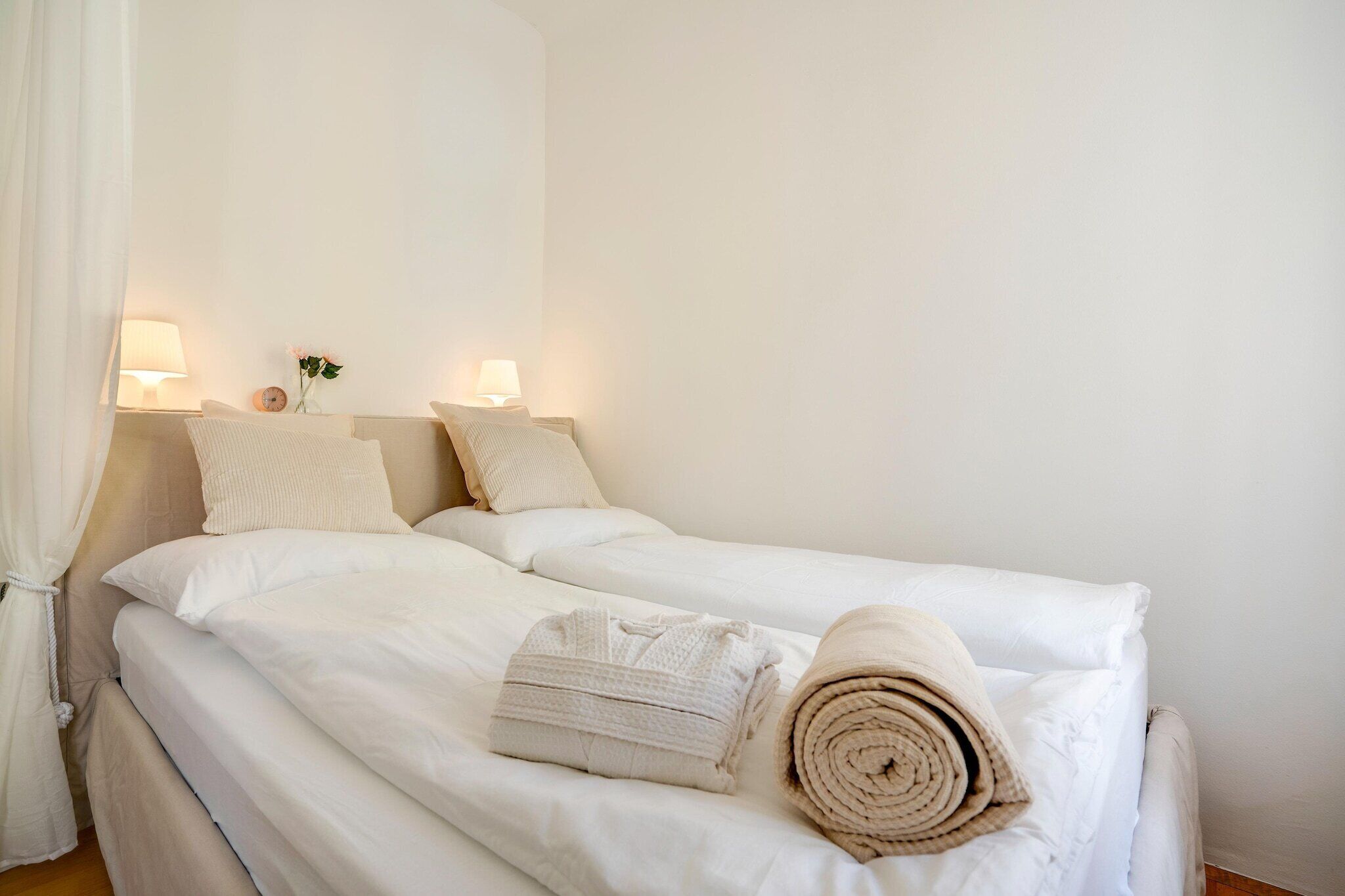 Ein Hotelzimmer mit zwei Betten, weißen Laken, einer Nachttischlampe, einer Vase mit Blumen und zusammengerollten Handtüchern.