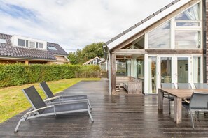Ein modernes Haus mit einer Holzterrasse, Sitzgelegenheiten im Freien und großen Fenstern.