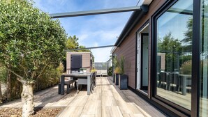 A modern outdoor patio with a dining table and chairs, surrounded by a wooden deck and a glass enclosure. - Modern Chalet near Beach in Renesse (Renesse)