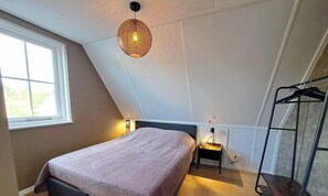 A bedroom with a sloped ceiling, a bed, a hanging light fixture, a nightstand, and a coat rack.