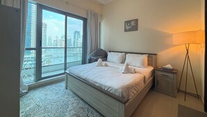 A hotel room with a large bed, a nightstand, a lamp, a view of the city through a large window, and a painting on the wall.