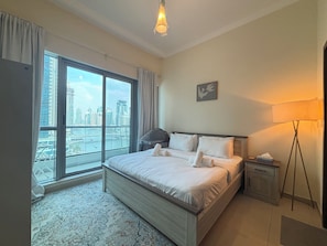 A hotel room with a large bed, a nightstand, a lamp, a view of the city through a large window, and a painting on the wall.